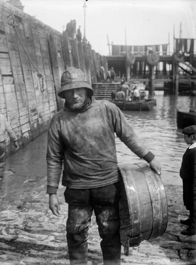 Detail of A Folkestone fisherman in smock and sou'wester carrying a tub for fish on the slipway by unknown